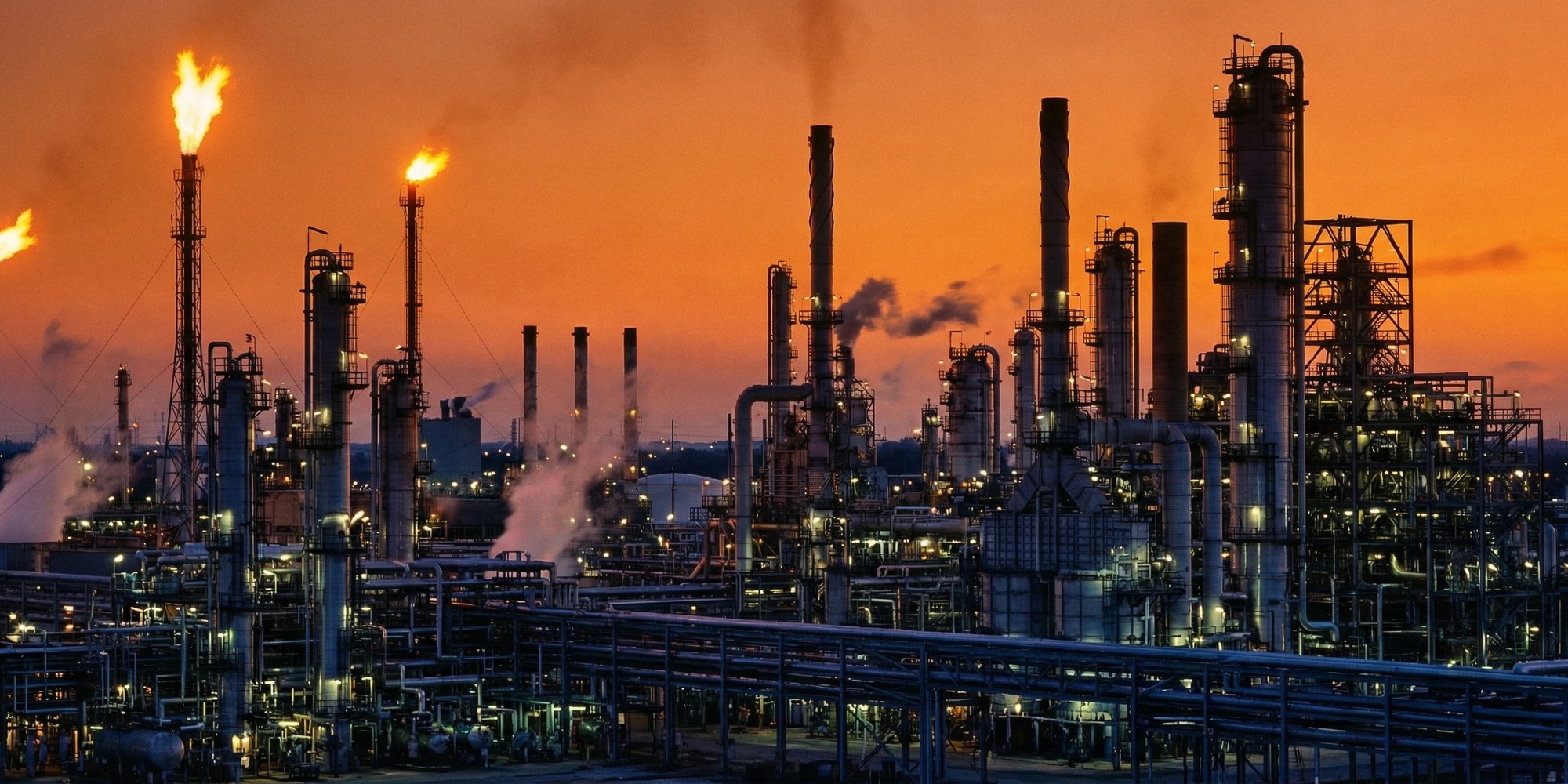 Oil refinery infrastructure at dusk, industrial petroleum processing facility with glowing flare stacks and complex pipe networks against an amber sky