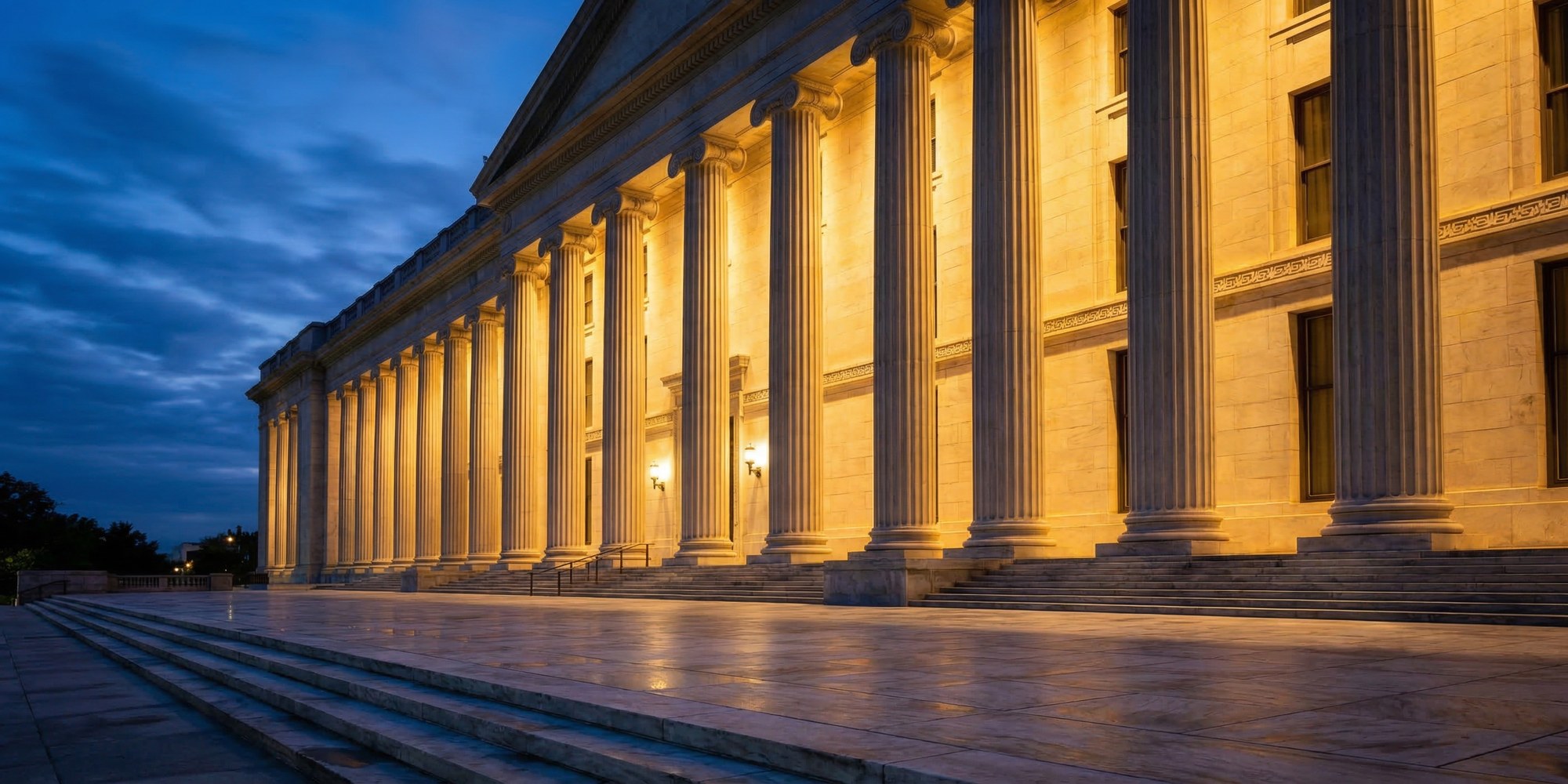 Federal Reserve Eccles Building in Washington D.C. at dusk, neoclassical limestone columns illuminated in warm amber light with empty marble plaza in the foreground
